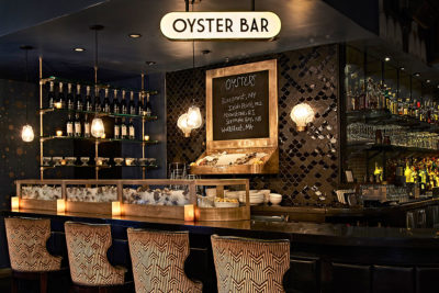 The oyster bar at Roxy Hotel. There are shelves of liquor bottles, a menu board and oysters over ice on display.