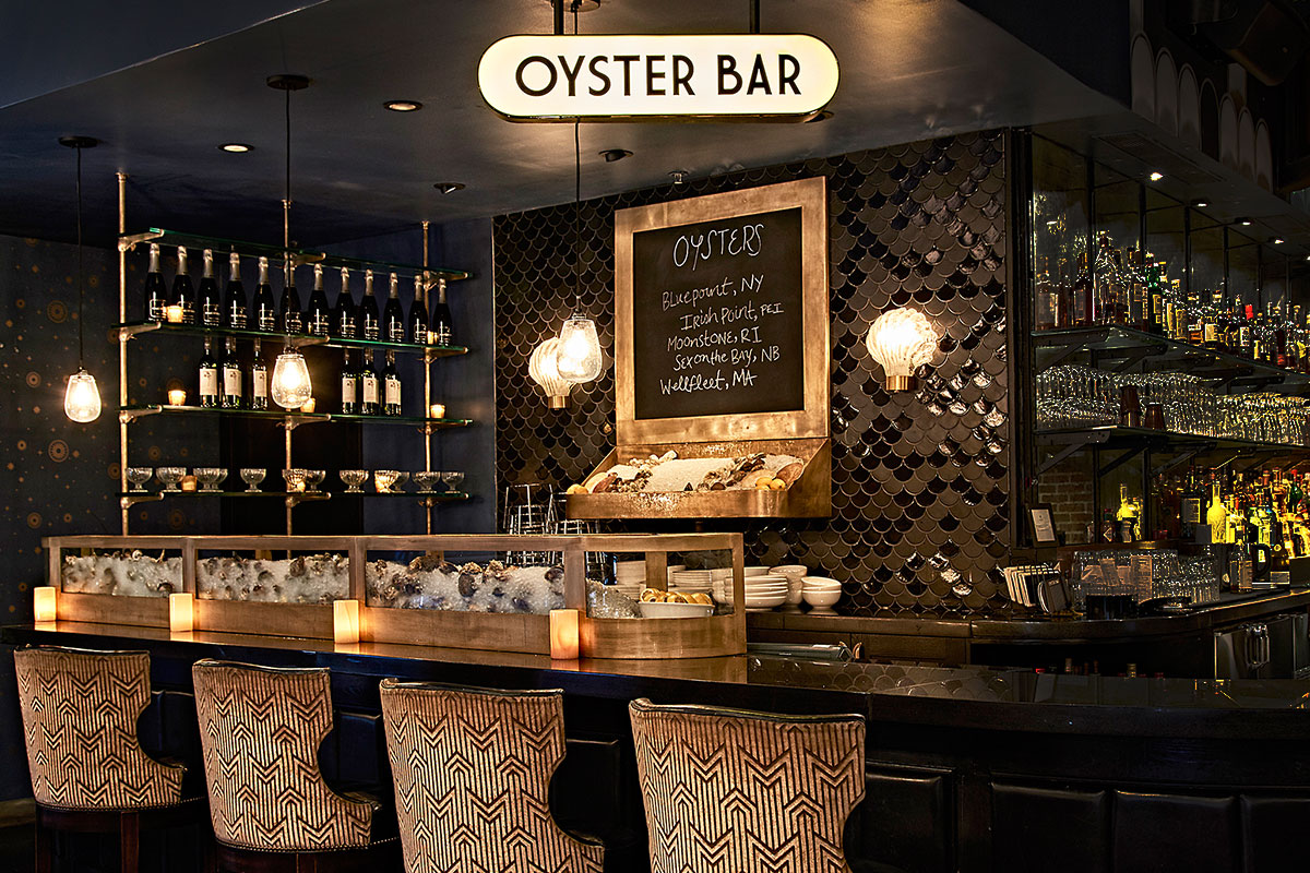 The oyster bar at Roxy Hotel. There are shelves of liquor bottles, a menu board and oysters over ice on display.