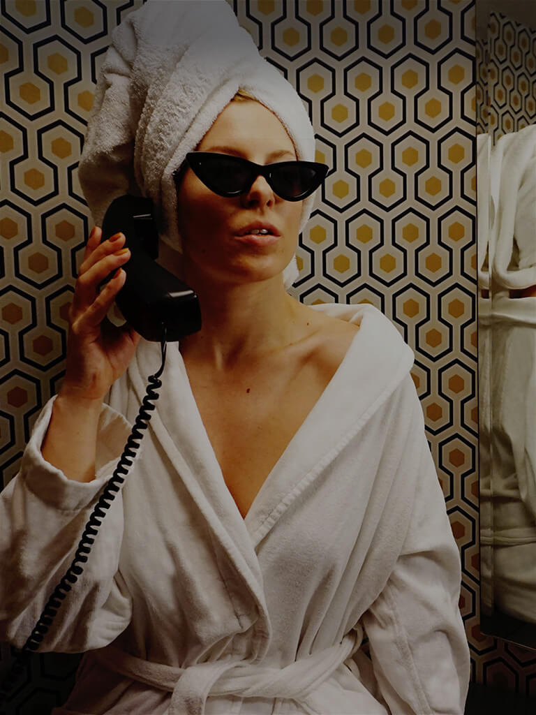 Woman in a white robe and sunglasses talks on a retro phone in a bathroom at Roxy Hotel.