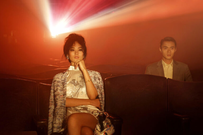 A chic woman sitting in a movie theater, backlit by the movie projector, with a man sitting a few seats away.