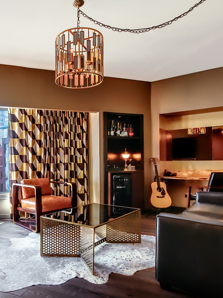 Living room of a deluxe one bedroom suite at The Roxy Hotel featuring a black leather couch, glass and metal table, gold chandelier, and hardwood floors.